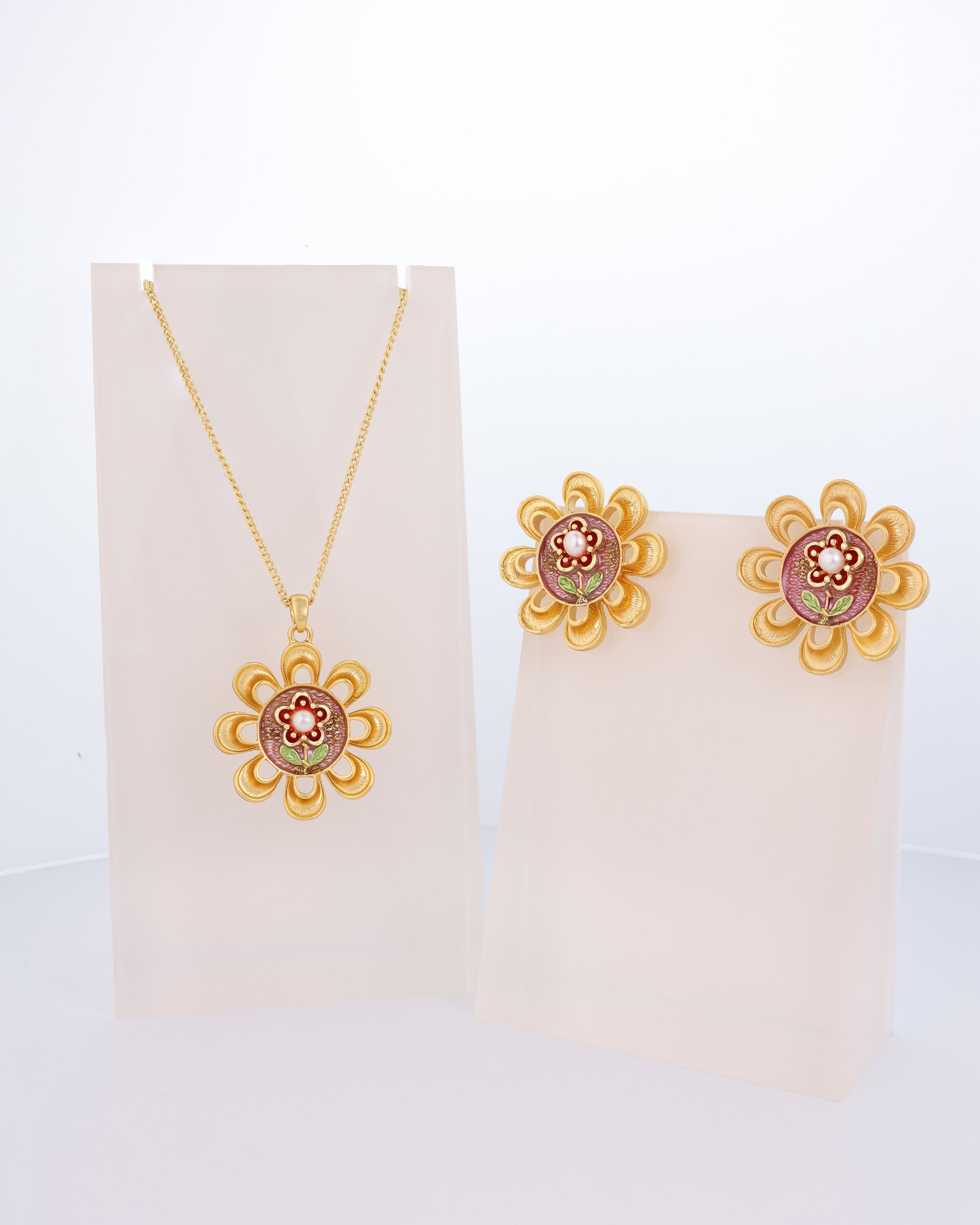 A gold-plated pendant necklace and earrings set featuring a floral design with red inlays on a white display stand.