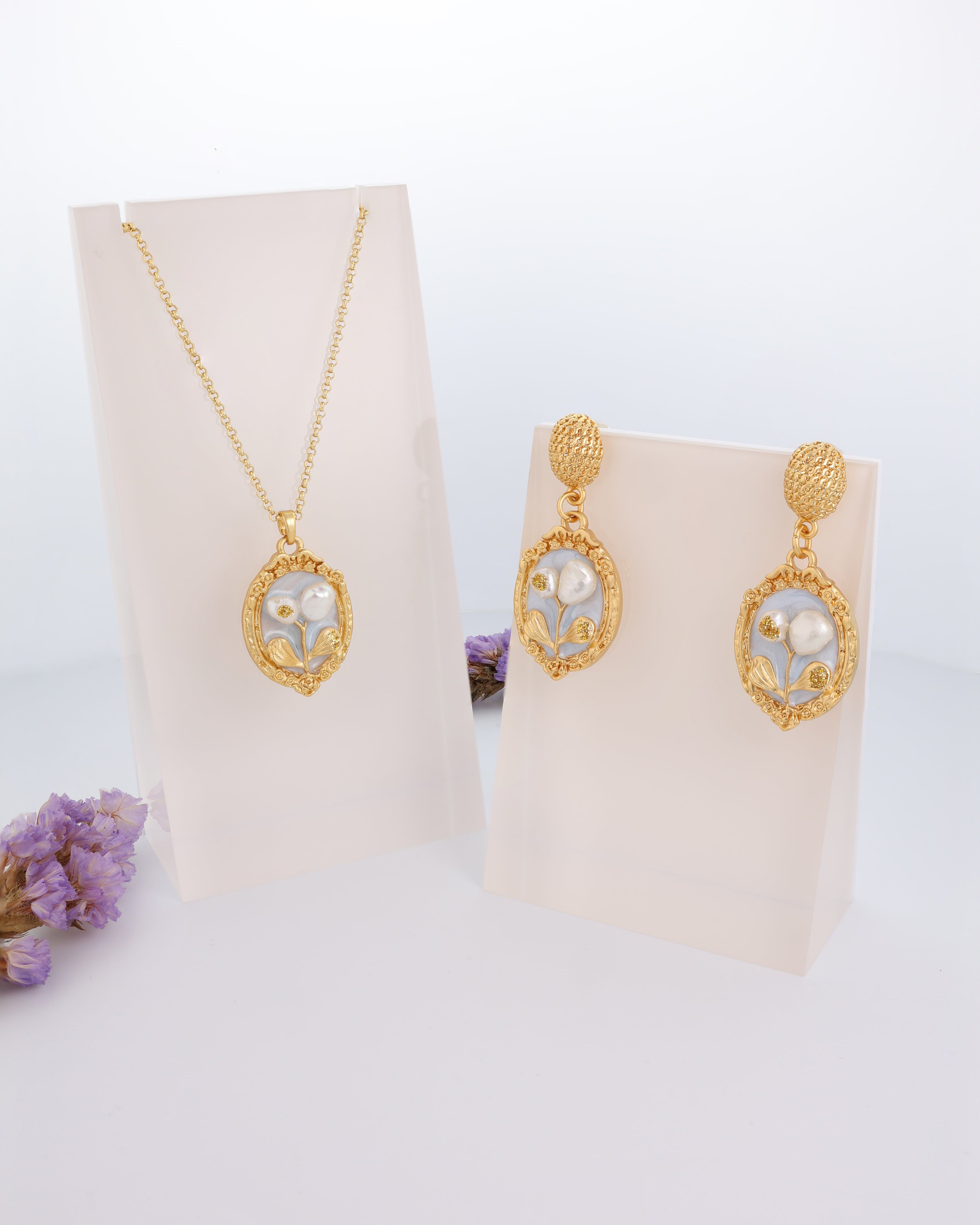 A pendant set displayed on a stand, consisting of a gold-colored necklace with an oval inlay containing a natural pearl, and a pair of matching earrings.