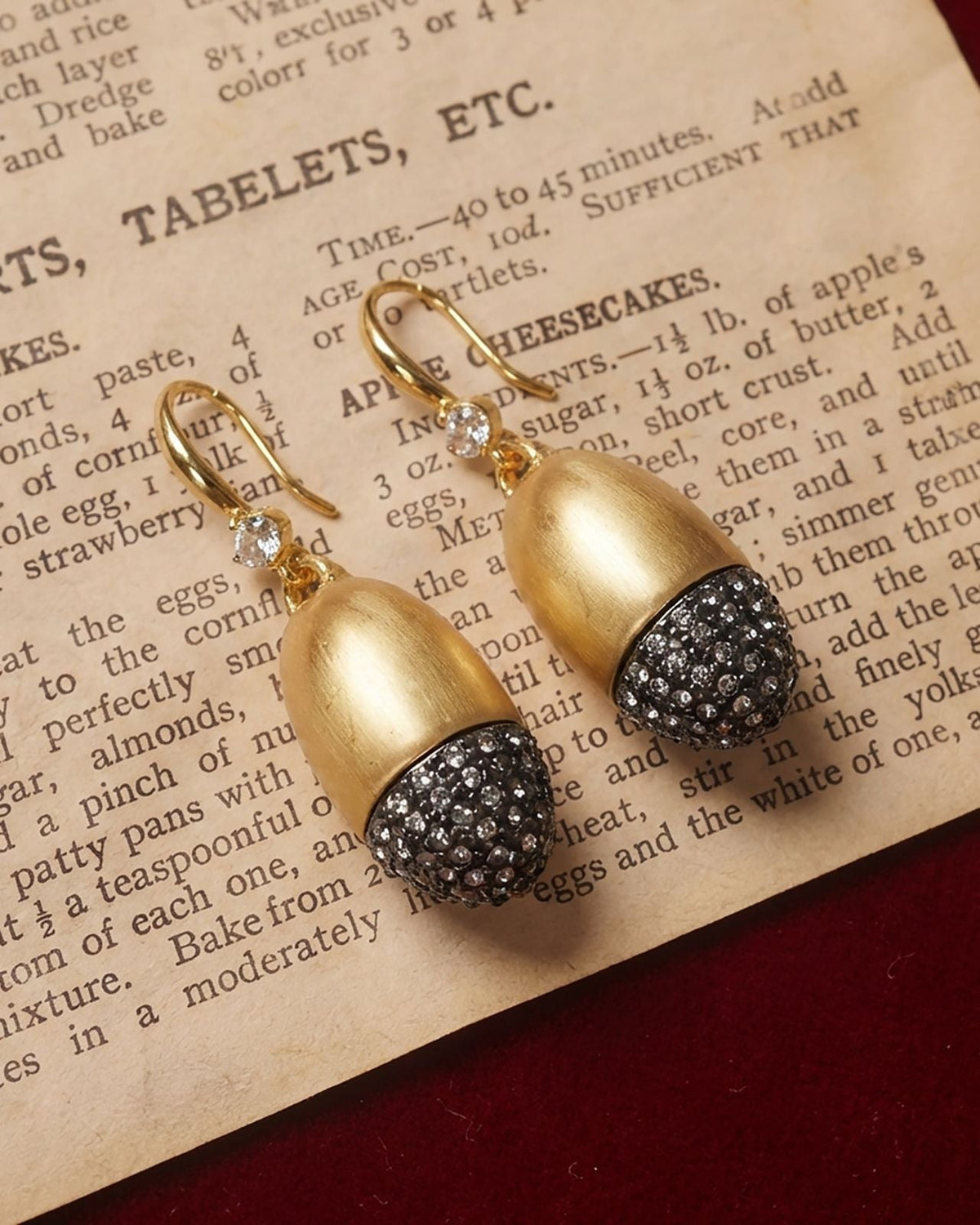 A pair of gold drop earrings with acorn-shaped charms, featuring a textured gold top and a black pavé-set crystal lower section, resting on an antique book page.
