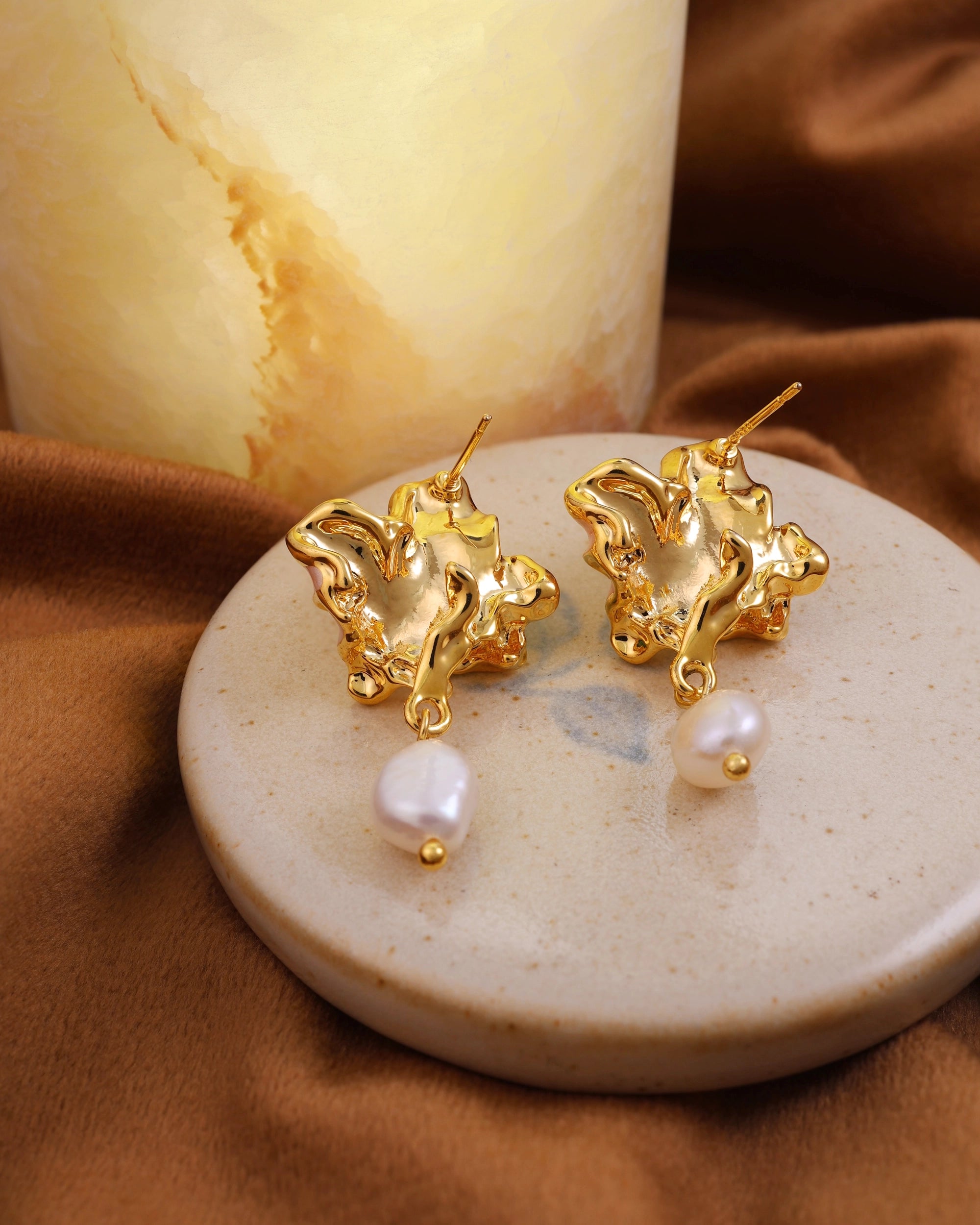 Gold earrings with pearl drops on a beige plate against a warm-toned fabric background