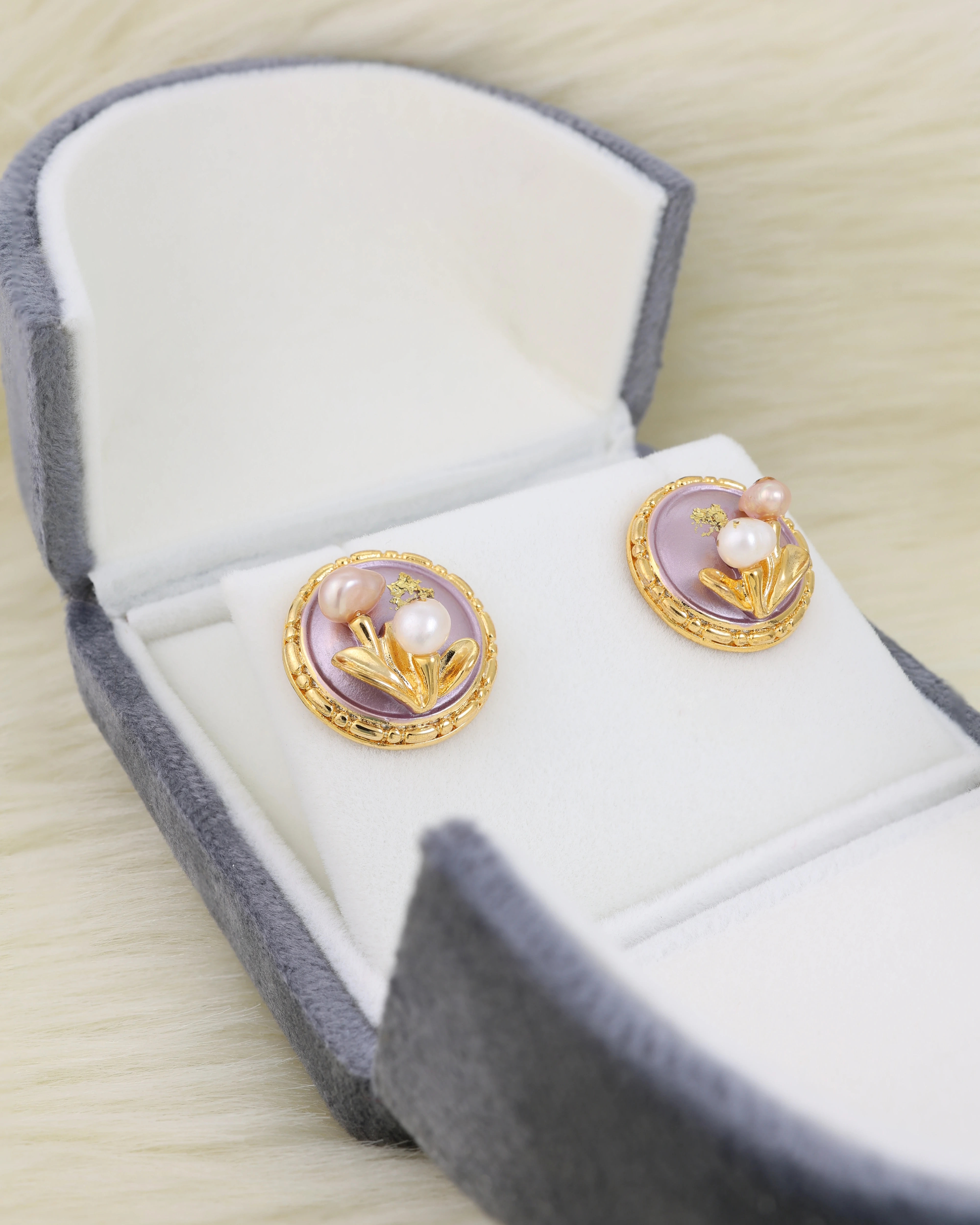 Pair of earrings with purple and gold design in an open jewelry box on a light fur surface.