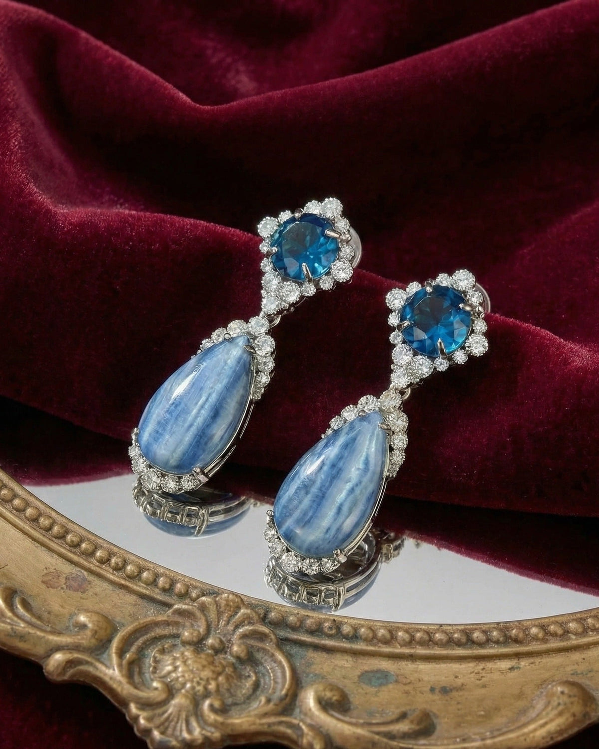 A pair of drop earrings with blue teardrop-shaped stones and blue oval stones surrounded by small clear crystals, displayed on a reflective surface with deep burgundy velvet fabric and an ornate gold frame behind them.