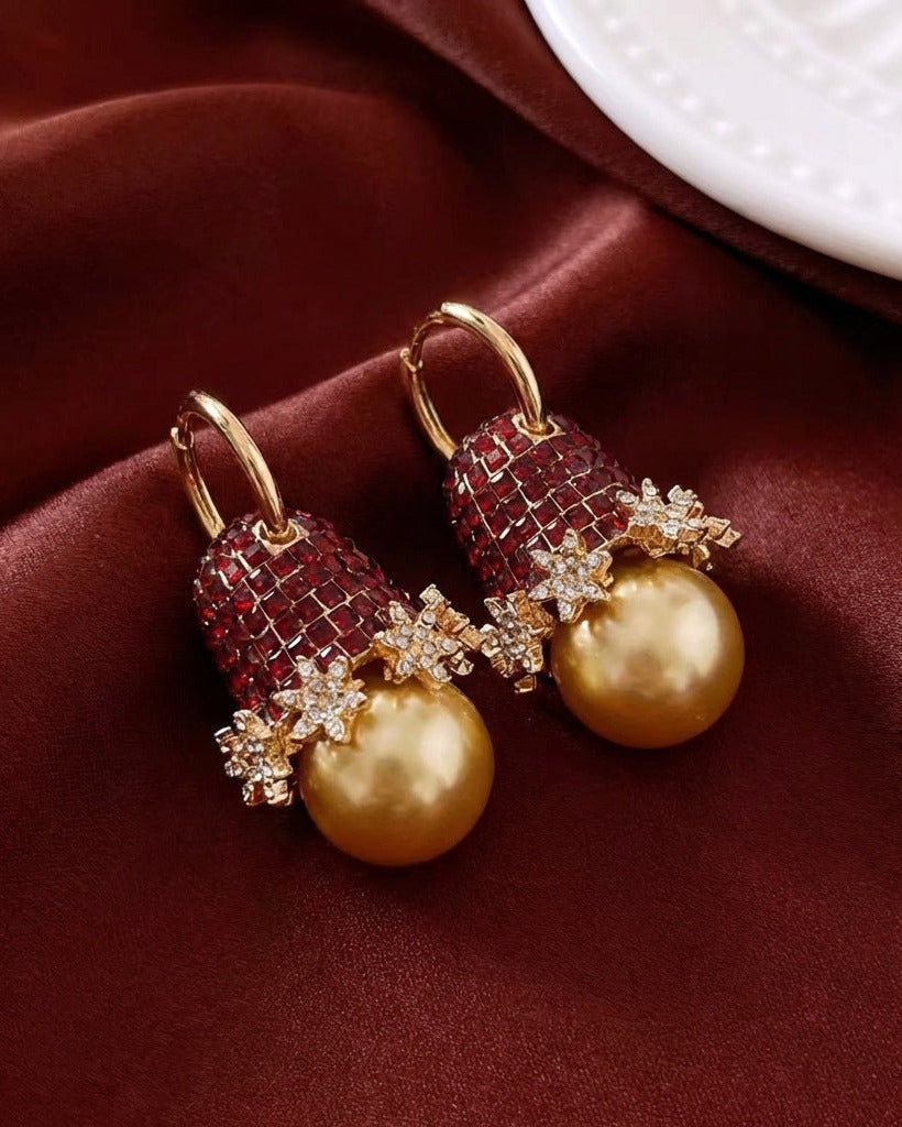 A pair of gold hoop earrings with textured red and gold drop charms, featuring a large gold pearl and crystal snowflake details, displayed on a dark red satin fabric.